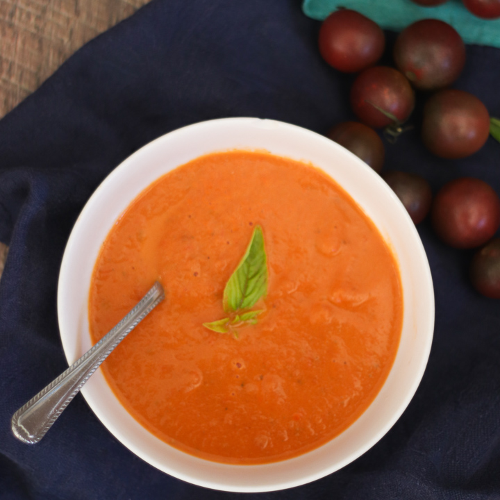 tomato soup in bowl with spoon and sprig of basil; blue cloth under bowl and cherry tomatoes in background